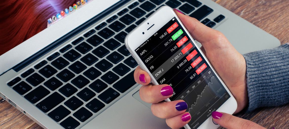 Woman with purple nail polish looking at stock market figures on mobile device with laptop in background